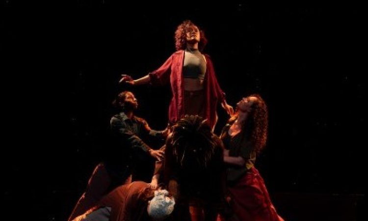 A dancer stands atop a group of other dancers, expanding their chest and focus upwards with an uplifted energy. They wear a maroon button-up shirt and don curly grown hair. The other dancers bend in towards this dancer, supporting and lifting them up. They wear multicolored pedestrian clothing and are lit by a warm, dim, light. They are soft with energy that extends upwards.