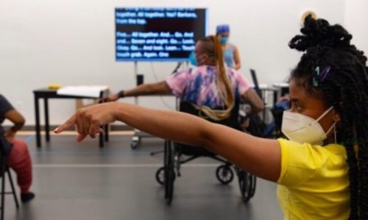 How We Move artists rehearsing majorette dance in the studio. Featured are Hector Machado, Jackie Robinson, and Kayla Hamilton. Everyone is masked with their left arm and index finger pointing outward to the side, and their right hand on their hip. The shot is taken from behind the dancers. In the front, captions are being shared on a large TV screen.