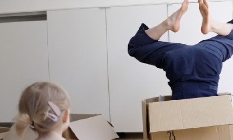 Selena Rook stands in a white-walled room wearing a sleeveless navy blue jumpsuit. Perched on her right hip is a medium sized cardboard box.