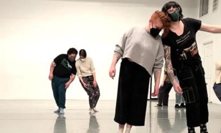 In a bright white room with light grey marley, dancers are clustered to the right side of the image in pairs, sharing weight. Two pairs of dancers are connected at the shoulders, one pair is connected by the hands, and we can see the legs of two hidden dancers standing back-to-back. There is an implication of motion, through shadows, blurred limbs, and raised feet.