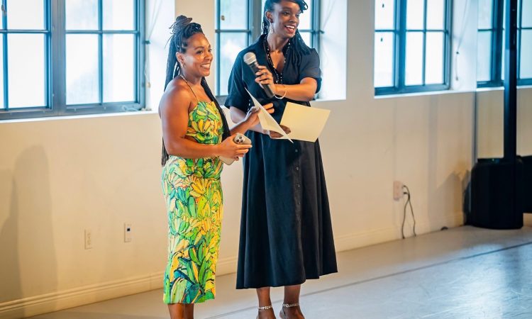 The co-founders of Banyan Dance Festival are two black women, Branndi Lewis and Ericka Squire. They stand in a dance studio, wearing cocktail attire, a green fitted dress for Lewis and a black button-down collared dress for Squire. With glittering smiles, they share a microphone and hold paper notes for their address to the intimate audience gathered at Ballet Florida for the Banyan Dance Festival gala and preview evening taking place the night before the mainstage performance.