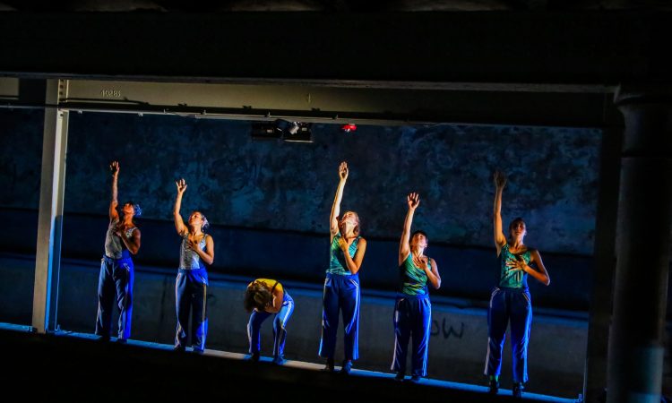 The six female dancers of The Naked Stark stand in a line facing upon a raised ledge above the subterranean Kelly Pool (not visible to the viewer). They face forward, wearing bright, electric green, patterned swimsuits and blue sweatpants. Five of the women gesture with their right arms stretched straight upward with their bent left arms resting on their chests. Each looks up toward her extended arm. The sixth woman forward bends over, her head and face into her torso, hiding. Precise side lighting illuminates their bodies with a silvery glow in the otherwise dark space.
