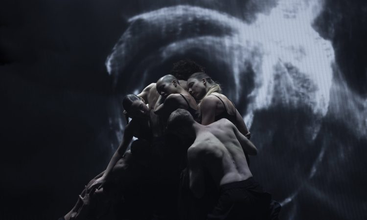 A group of dancers stands in a tight cluster, in shadowy lighting. A black and white video projection lights up the backdrop behind them.