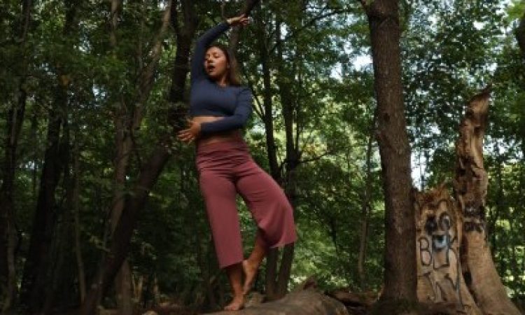 Balanced on a fallen tree in a wooded area a dancer poses wearing maroon pants and a blue long sleeved crop top. She balances on her right foot, touching her pointed left foot against the right ankle. Her left hand reaches across her midsection with the hand and fingers spread wide. Her right arm reaches up over her head, fingers spread wide, palm down. She softly gazes down, her mouth relaxed open.