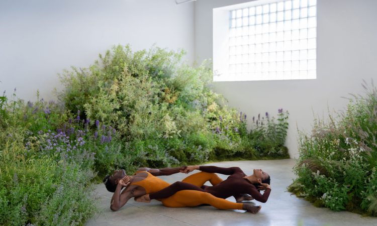 Lloyd Knight and Jacquelin Harris, both Black dancers, are on the ground in a gallery with white walls, a large glass block window, and a gray cement floor. An installation of wildflowers and grasses surrounds them. Knight wears a copper sleeveless unitard and Harris a brown long-sleeved unitard. Their legs are intertwined, and their backs are almost touching the floor. Their faces gaze up at the ceiling, eyes closed.