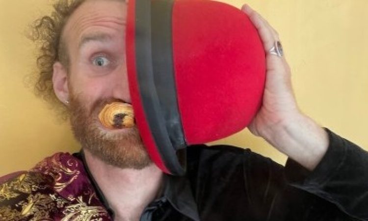 A white person with curly hair, a beard, and piercing blue eyes shows half of zir face, covering the rest with a red dome shaped hat. Pain au chocolat is stuffed in zir mouth, and zir clothes are bifurcated, much like zir face––half outfitted in red and gold, and the other half in black.