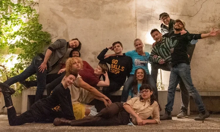 A group of twelve people pose in front of a white wall. on the floor level, four group members either sit, lie, kneel or lunge on the ground. standing members lean on one another on either side of the crew, and people in the center sit or lay on a bench together.