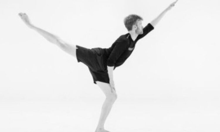 A young man facing the right, wearing black shorts and matching black t-shirt, beautifully extends his left arm forward and right leg back.