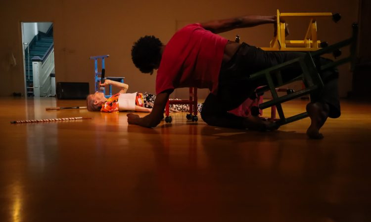 In the foreground and in darkness, dancer Zo Williams is balanced on his left forearm and left shin. In between his legs is a tiny wooden chair, beneath. another yellow one he holds in place with his right hand. Zo faces upstage, where Cathy Weis lies on the floor on her back, with what looks like two canes lying on the floor on either side of her head. More brightly illuminated, Cathy holds a microphone up to her mouth, a tiny blue chair beyond her. In the distance, through an open door to the hallway, we see stairs going up.