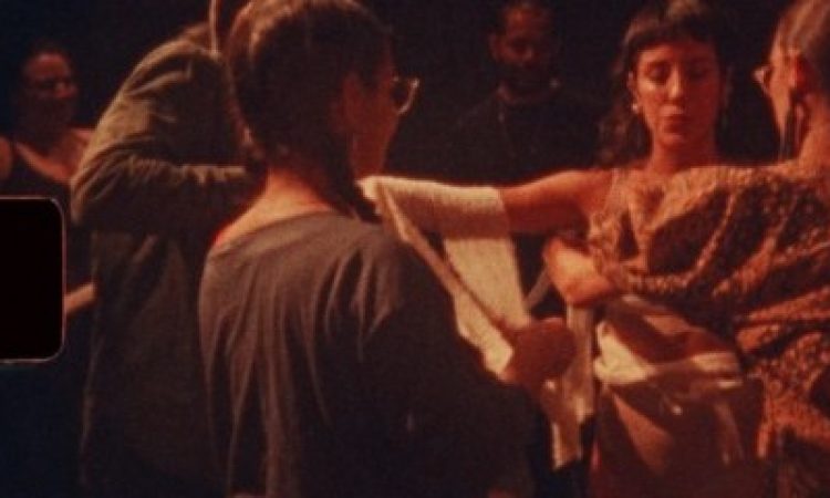 A scene from Ceremonia I: In Defense of Tenderness. Travieso, draped in white fabric with eyes closed in a somber, meditative expression, is surrounded by five audience members who look on, some gently adding cloth to her body. The image itself is shot on a 8mm film camera, lending a warm, faded grain that adds a reflective quality to the photo.