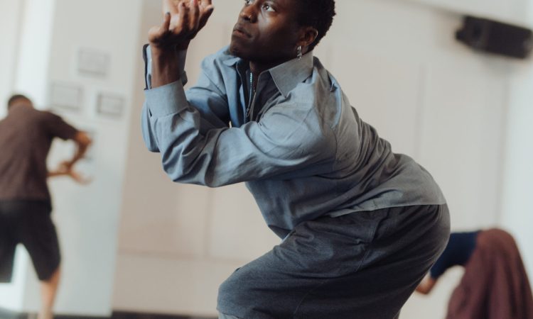A dancer wears layered gray clothing as they bend forward, somewhere between a crouch and a lunge. They push their palms out in front of their face, with fingers gently curved, as if pushing something upward into the air.