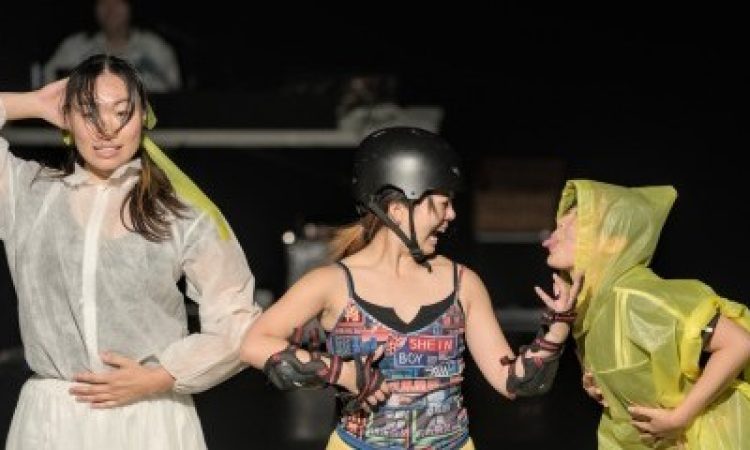 3 performers stand onstage–the first in an all-white mesh skirt and tulle skirt, blankly looking directly at the camera with a forced smile, one hand to the head, and the other on her stomach. To her right, two performers mingle, teasing each-other and laughing. One is outfitted in protective gear–a helmet, kneepad, elbow pads–and a multicolored SHEIN shirt with yellow bermuda shorts. The other is in a yellow poncho, gleefully sticking her tongue out, taunting her companion.