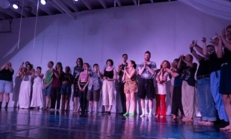 Full cast clapping onstage with the house lights up in a blue and purple light, taking from a low angle shot to their right.