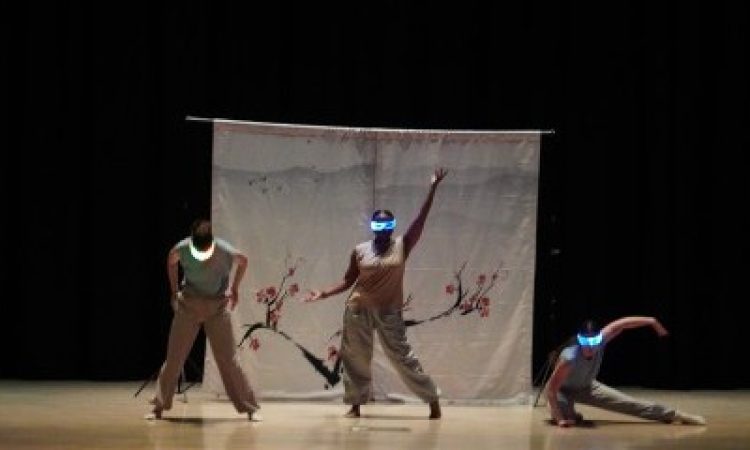 Five dancers on a wooden stage wear bright, glowing glasses. They all strike a different pose - some lift their arms and stare forward, while others reach to the side and lunge. Behind them is medium-sized white tapestry depicting a cherry blossom tree and behind that: a floor-to-ceiling length black curtain.