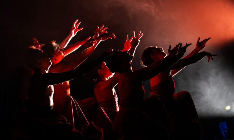 A group of shadowy-figured dancers in red kneel in profile. They reach their fingers toward a moody, red light in the upper right hand corner. The whole scene is bathed in haze