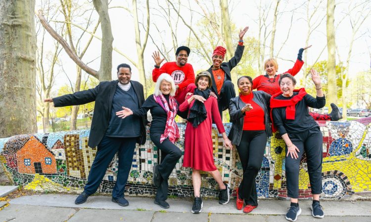Eight senior dancers pose in front of a mosaic wall and a park. Their smiles wide, they open their arms, wave, and one points towards the camera. They are wearing black and red.