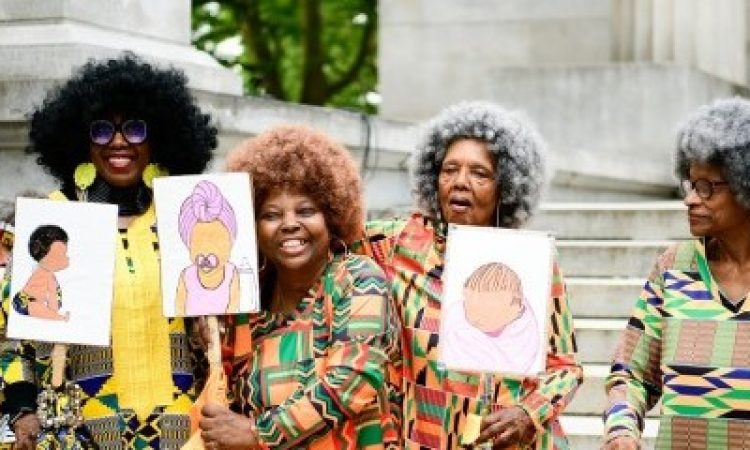 Standing in front of an old building with stone steps, four Black women of various ages stand wearing vivid shirts with complex patterns. All donning afros, three of the women hold up signs which show hand-drawn illustrations of infants or children, while the fourth has her hands resting on her walker. Two of the women smile radiantly.