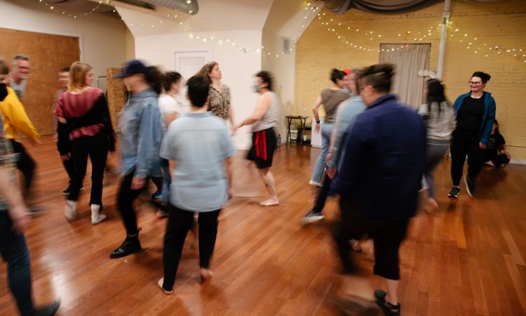 A crowd of audience participants walks through a warm room with a wood floor and string lights in the background. Mid-motion, the camera blurs most of the participants as they smile and walk in-between and around each other.