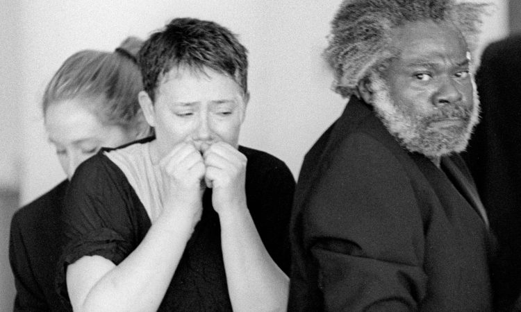 Three people are in this black and white image. The center person looks down, their hands in fists, scrunched up to their lips. Another man with a beard, wearing a suit jackets, stares directly into the camera. The last person is partially obscured, looking down.