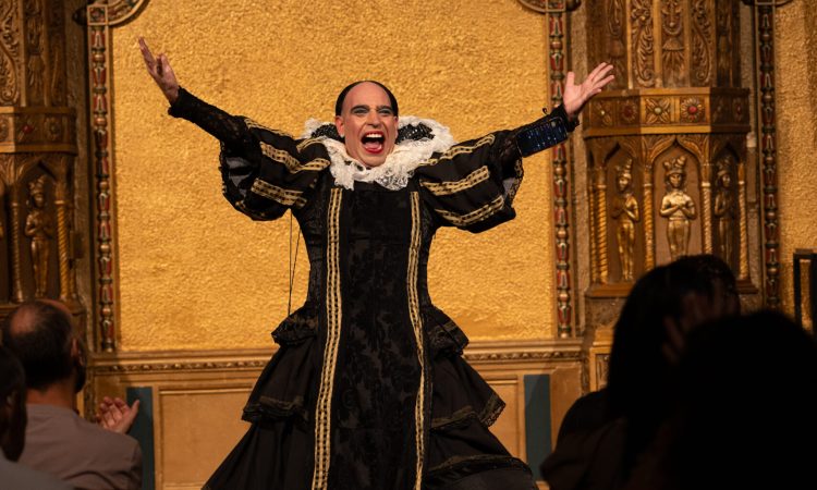 Edu Dìaz is dressed in a black gown with a large white lacy collar center on a golden gilded stage. Their arms are up exubrantly akimbo.
