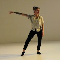 Emilee Lord stands with her weight on her left hip and her right arm extended to the side of her body. She wears a loose shirt and black pants and socks.