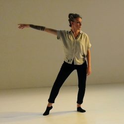 Emilee Lord stands with her weight on her left hip and her right arm extended to the side of her body. She wears a loose shirt and black pants and socks.