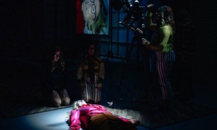 On a dark stage, a person lies supine on the ground. Two people kneel near her head, looking down. Above them all, a camera is pointed down. Behind the people is a projection of it all, visible and blown up for the audience.