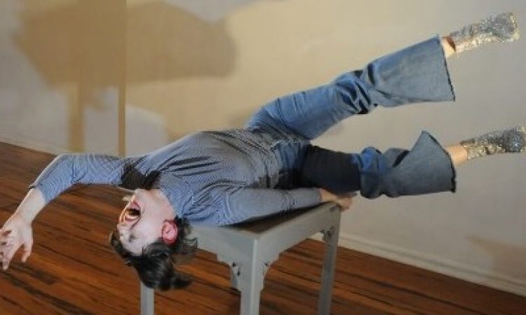 A white woman with dark brown hair, wearing a blue outfit, sparkly shoes, and a bright red lip that matches her red hoop earrings, lies back on a small gray table. Her mouth is wide open as if singing, one arm reaching emphatically above her head while the other grips the table tightly for balance. Her legs float in the air, parallel to one another at a slightly awkward angle. She is a rockstar in an empty room.