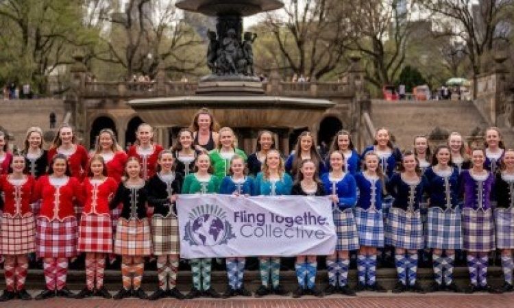 Two rows of Highland dancers stand in front of the Bethesda Fountain at one of the entrances to Central Park in NYC. There are trees in the background. The dancers are clad in bright plaids and knee socks in orange, red, green, blue, and purple. A few in the first row hold a sign that says Fling Together Collective