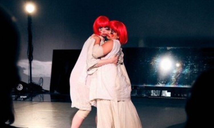 Two performers (Doan and Stapleton) stand cheek-to-cheek as they stare in confusion at the audience. They wear bright cherry-red wigs and garishly applied makeup along with white baby-doll overshirts. Behind them is a long mirror; a flash goes off.