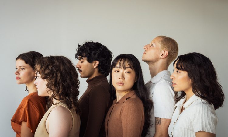 Five dancers stand in side profile, all looking in different directions and wearing earthy-colored cotton tops. Their faces are impassive and neutral.