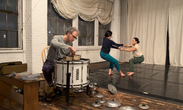 In the foreground, a percussionist, Flandrew Fleisenberg, wearing a grey long sleeve and glasses leans over and rings a brass soundbowl that he’s placed on top of a big white YAMAHA drum. Metal kitchen pot lids are strewn on the faded wood floor around him. In the background, a person in teal pants and a navy longsleeve, Loren Groenendaal, crosses arms and holds hands with a person wearing green pants and an off white t-shirt, Michele Tantoco. They counterbalance, sending their hips back and holding each other's gaze. Beneath their blurring feat, black marley crinkles, behind and beside them, white curtains drape the backstage and the windows.