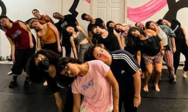 A large group of dancers stand closely together in a studio, their bodies tilted in unison with heads falling dramatically to one side, creating a strong visual wave across the room. They are arranged in staggered rows that draw the eye from front to back, emphasizing their collective rhythm and connection. The black floor grounds the image, while the wall behind them bursts with bold, curving black outlines and sweeping pink brushstrokes, giving the space an energetic, almost theatrical backdrop.