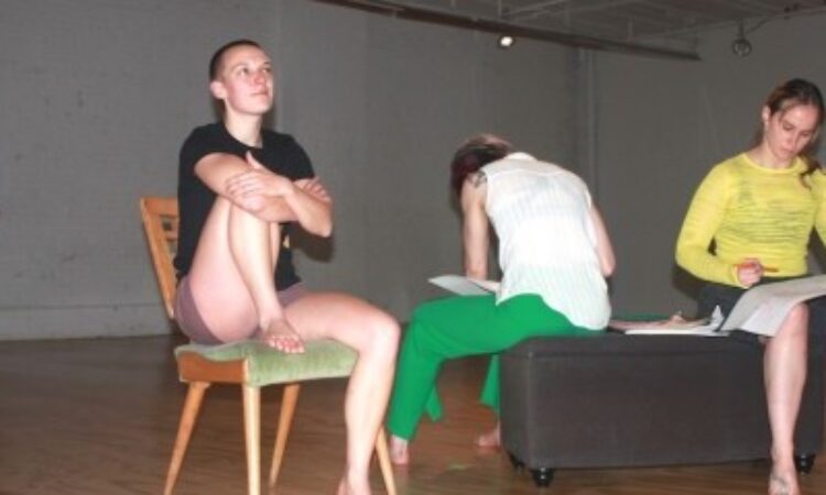 Three performers sit in the foreground of the picture: one resting with a knee up on a chair, gazing up into the distance with a slight smile; the other two turned at various angles (one facing us, one away) on an ottoman, drawing and concentrating on large sheets of white paper. In the background, dimly lit, a fourth performer stands, slightly bent, in a gesture of reaching.