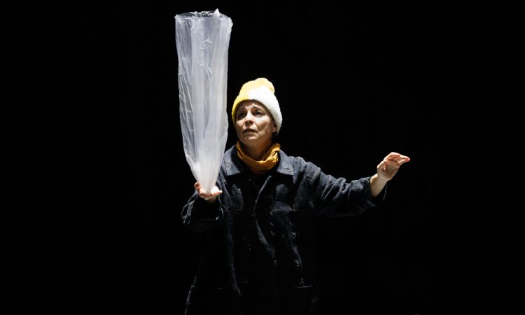 Under a spotlight in an otherwise darkened space, a woman in a yellow and white pompom knit hat, yellow scarf and black jacket balances a plastic bag in her right palm. She looks at it, brow furrowed. Her left hand is lifted chest high, palm facing down, a gentle curve in her closed fingers.