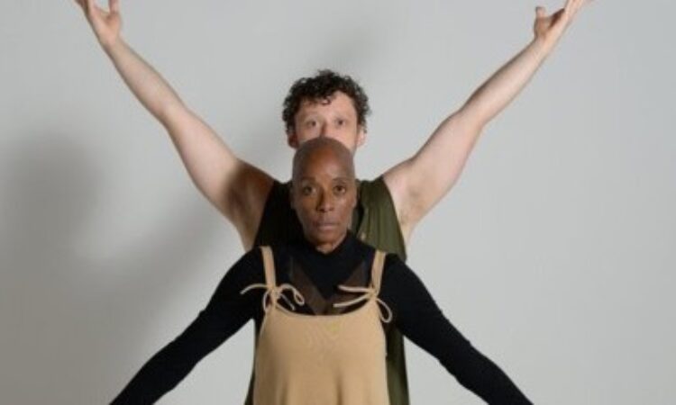 Rhonda Moore stands with her arms outstretched in a low “V” position looking directly at the camera. Ben Grinberg stands behind her with his arms outstretched in a high “V” position looking at the camera. Grinberg wears a green sleeveless top, and Moore is wearing a longsleeve black button-down under a light-brown jumpsuit. They stand in front of a gray backdrop.