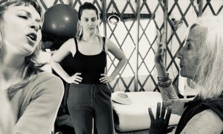 On the left, a young woman twists to her right, her arms bent and her mouth open, while on the right, a much older woman gazes at her steadily, with her bent arms up. In the center, watching the two of them work, stands another young woman with her hands on her hips.