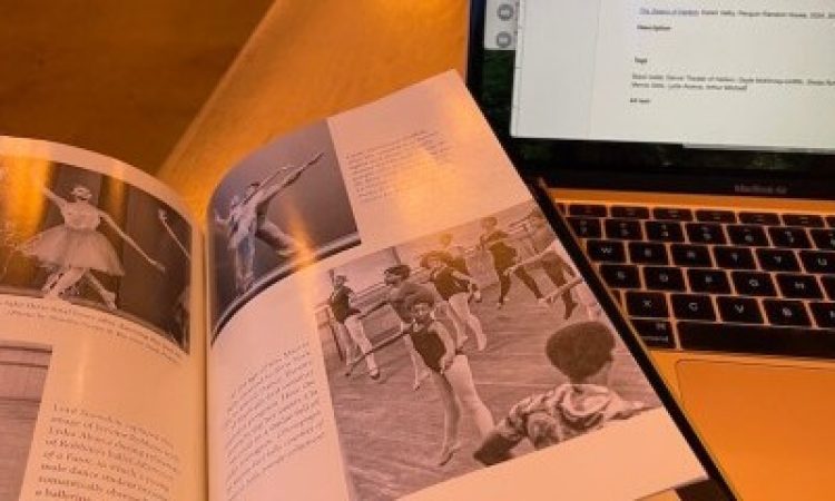 A copy of The Swans of Harlem is open on a table next to an open laptop. The book is open to a photo spread. The black-and-white images depict dancers from the early days of Dance Theatre of Harlem, including an image of Lydia Abarca being coached in Afternoon of a Faun, final bows, Gayle McKinney-Griffith being partnered in arabesque, and a photo of Marcia Sells in class at ten years old. Sells is jumping in second position with her feet pointed and arms outstretched, looking down towards her left hand.
