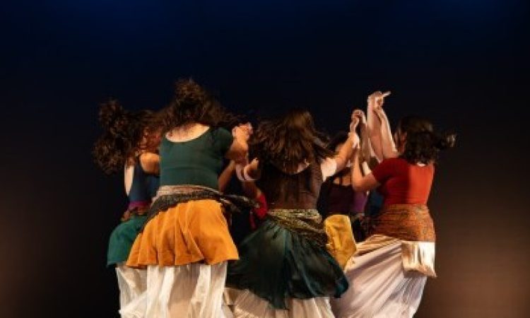 Six dancers come together in a circle. Their hair and their long white skirts flare with movement as their grasped hands reach in and up, towards the center of their dancing circle.