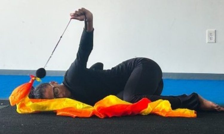 Performer Daisie Cardona rests in a fetal position on her side with her eyes closed. Dressed all in black, one arm stretches above her body, holding a black string taut. The string is connected to a red, orange, and yellow ribbon which rests on the ground.