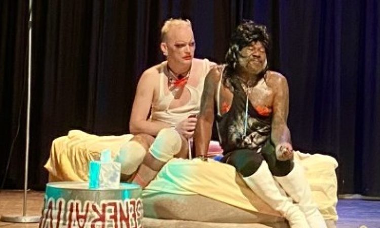 Two queer people sit on an inflatable bed. A metal basin flipped upside down is used as a table.