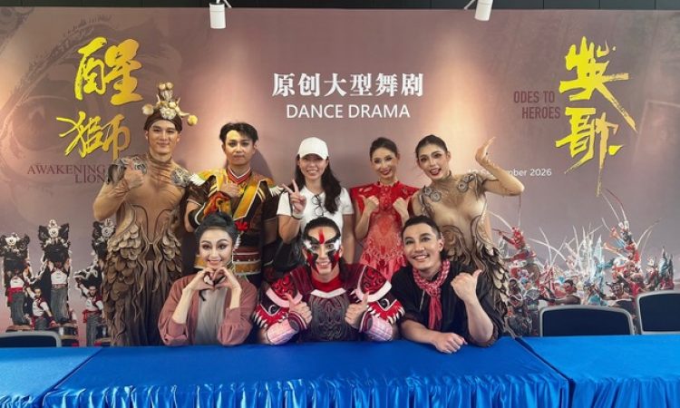 The author, Ziying Cui, wearing a white cap and T-shirt, stands at the center of seven Guangzhou Song and Dance Theatre dancers after a performance of Odes to Heroes. The dancers wear colorful costumes and stage makeup, smiling and posing with thumbs-up and heart-shaped hand gestures behind a blue-covered table. A backdrop displays the titles and imagery of both productions.
