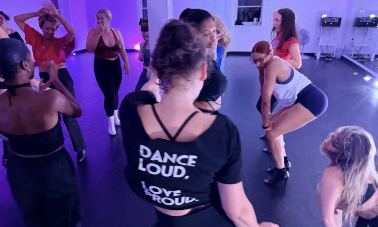 Overhead view of a dance class forming a circle under purple studio lights. A dancer in the center foreground wears a black t-shirt reading "DANCE LOUD, LOVE PROUD" while others around her cheer, dance, and watch.