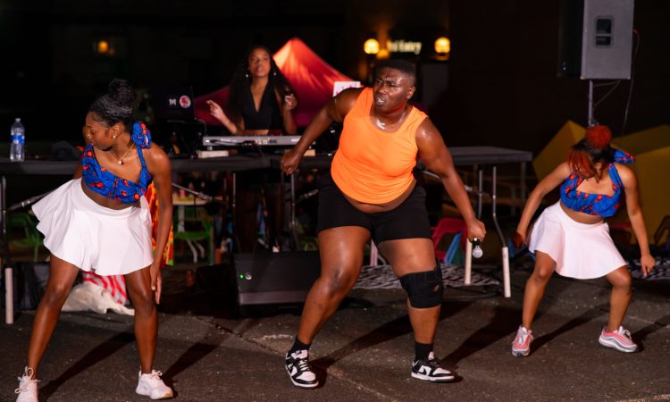 Bempa, wearing a bright orange shirt, black exercise shorts and sneakers, is framed by two black and brown femme dancers wearing matching blue and white outfits. They are dancing outside at night, lit by stage lights on a sidewalk with a femme appearing DJ playing music in behind them. The dancers have bent knees fronts facing the audience mid twist, looking over their shoulders with concentrated expressions.