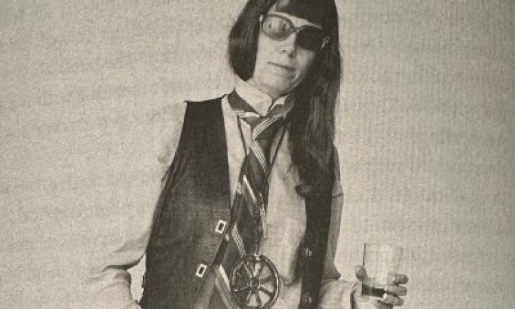 A photo of a page in the book. A black and white photo of Jill Johnston. She has long hair, aviator sunglasses, and is wearing a white long sleeve button up, a black vest, and a wide striped tie. The photo is cut off at her hips. She leans into her left hip and has her right hand in her pocket. A glass in her left hand.