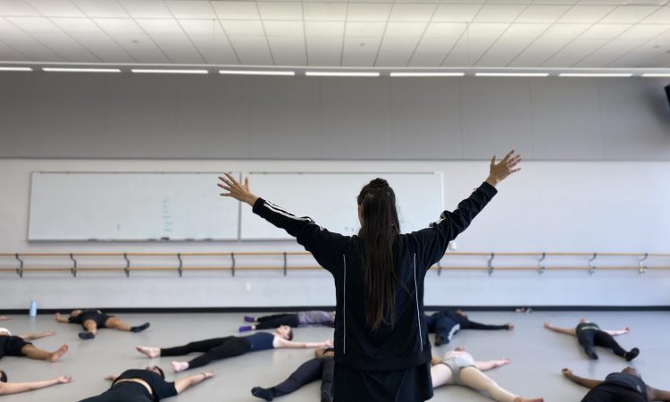 In a dance studio with white walls, a light gray marley floor, and two large whiteboards along the back wall, situated above a double-stacked wooden ballet barre, eleven college students lay supine with their arms raised and legs parted in an “X” shape on the ground. They lay at different angles, breathing deeply, following the instructions of the teacher at the front of the room. The instructor, tD writer Jennifer Passios, faces the students. She wears a black Adidas jacket. Her hair falls in a half-up-half down updo as her arms reach skyward in the same “X” gesture that the students make on the ground.