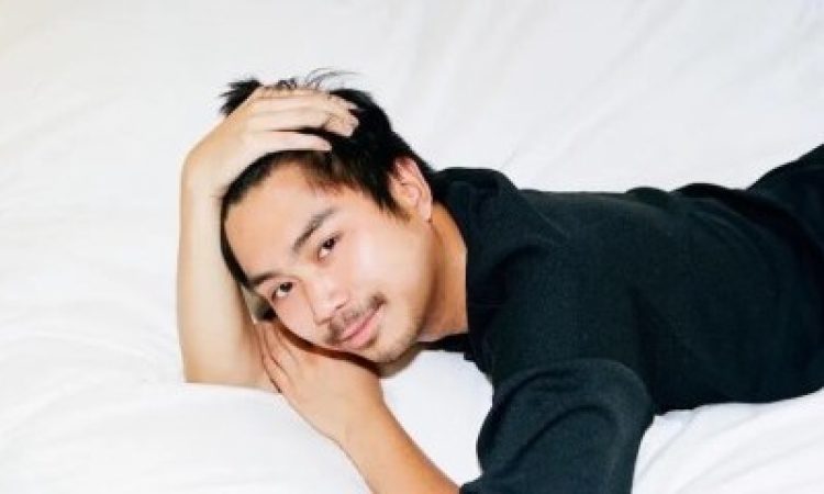 An Asian, male-presenting figure wearing black, lounges on plush white sheets, smiling as he frames his face with his hands.