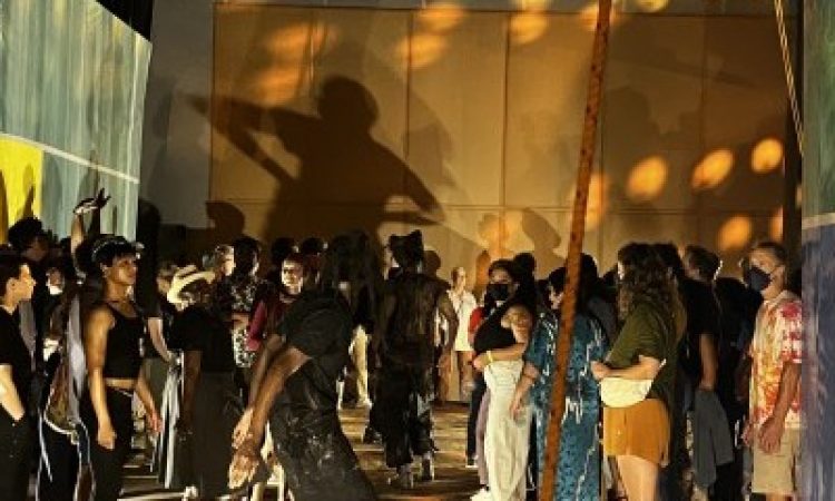 Inside a dimly lit warehouse type space, a crowd of onlookers surrounds 2-3 visible performing artists who wear all black. Green, blue and yellow paintings double as walls. A large shadow of one of the dancers looms above the crowd, projected onto the back wall in warm tones.