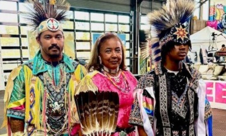 Vaughnda Hilton, her son Andrew Lyn, and Andrew’s son Jordan Lyn stand together—three generations of dancers. Hilton wears a jingle dress adorned with handmade beadwork and rows of metal (healing) cones–each cone represents a day of the year– while her son and grandson wear Grass Dance regalia featuring vibrant colors, intricate ribbonry and fringe, roach (headdress) and decorative elements that highlight traditional artistry.
