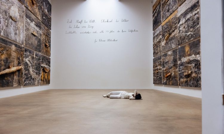 Dressed in white, performer Marguerite Hemmings lies prone on the floor of a large museum gallery at Mass MoCA between monumental earth-textured wall panels, beneath handwritten text on the far wall.